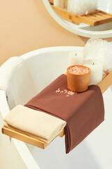 Close-up of a wooden tray placed across the bathtub with cloth towels, pink salt, scented candles and a bath sponge. Space with two main colors white and brown. Relax inside the bathroom.