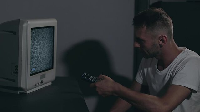 Broken Television or TV Propaganda. Mind Control - Zombified Man in White T-shirt Switches Channels on the Old TV in Dark Room. TV Signal Search. Distortion on the Screen. Medium Close-Up, Film Grain