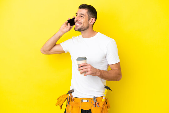 young electrician caucasian man isolated on yellow background holding coffee to take away and a mobile