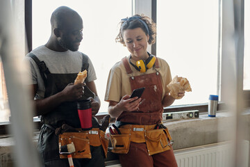 Smiling construction worker using smart phone and having lunch with coworker at site