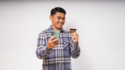 Portrait of satisfied happy Asian young man holding smart phone and credit card, entering data,...