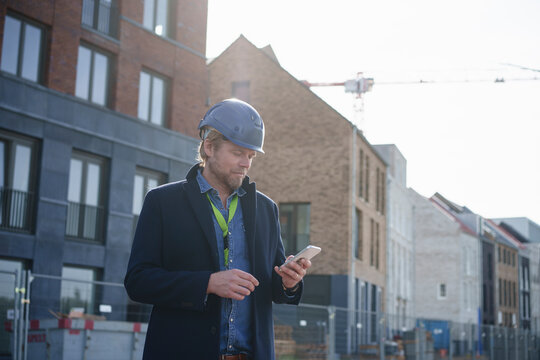 Architect wearing hardhat and using mobile phone near buildings