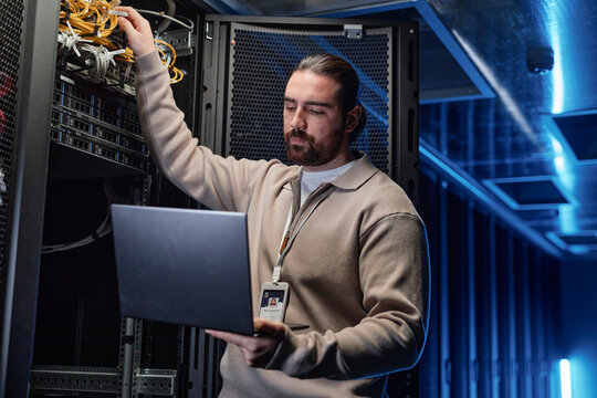 Young engineer touching cables and using laptop in server room