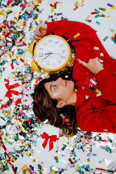 Smiling girl holding clock and lying on confetti