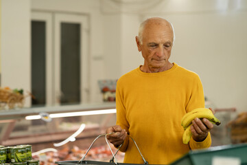 Fototapeta premium Old male buying food at supermarket, choosing bananas in fruit department. Grocery store shopping.
