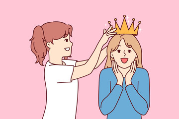 Little girl places crown on overjoyed sisters head while playing princesses from ancient kingdom. Two sisters from junior school try on golden crown received for achievements in education