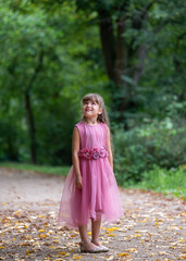 Portrait of a cute little girl 6 years old with long, dark hair. A smiling girl stands on a path with yellow leaves in the park in a beautiful pink dress and looks up with interest
