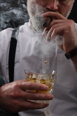 Bearded man with glass of whiskey smoking cigar against black background, closeup