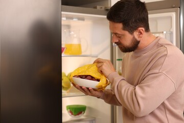 Man putting bowl covered with beeswax food wrap into refrigerator indoors