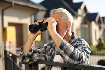 Fototapeta na wymiar Concept of private life. Curious senior man with binoculars spying on neighbours over fence outdoors