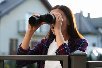 Concept of private life. Curious young woman with binoculars spying on neighbours over fence...
