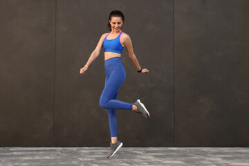 Beautiful woman in stylish sportswear jumping near black wall outdoors