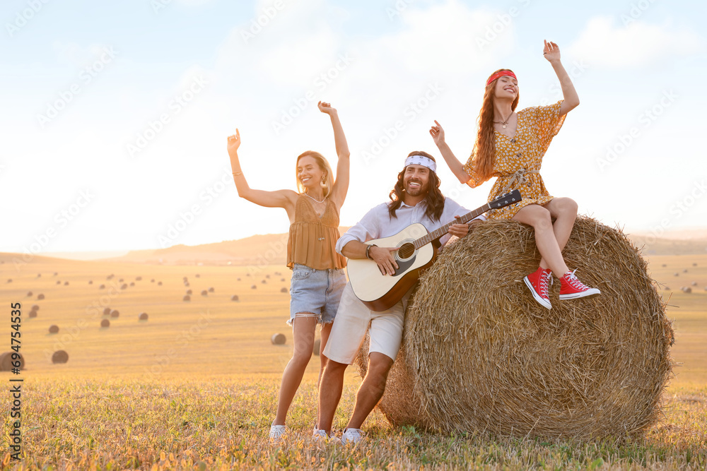 Sticker Beautiful hippie women listening to their friend playing guitar in field, space for text