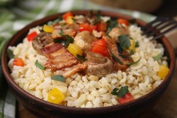 Cooked bulgur with vegetables, fried bacon and mushrooms in bowl on table, closeup