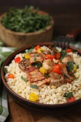 Cooked bulgur with vegetables, fried bacon and mushrooms in bowl on wooden table, closeup