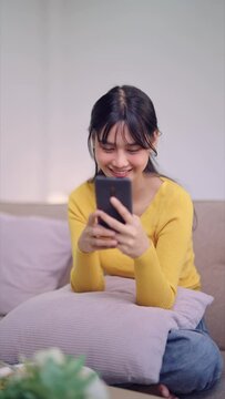 An Asian young woman chats on social media or video calls with friends on her smartphone, seated on her sofa, sipping coffee at home. Capture the cozy moments of virtual connection