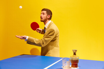 Handsome young man in elegant suit player table tennis against bright yellow background. Whiskey...