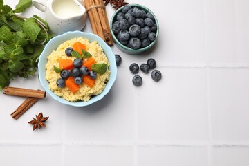 Tasty millet porridge with blueberries, pumpkin and mint in bowl on white tiled table, flat lay....