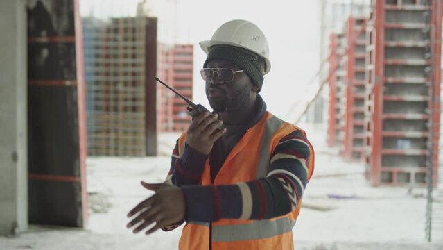 Medium shot of Black male foreman wearing reflective vest and hardhat using walkie talkie and making hand gestures while controlling work at construction site