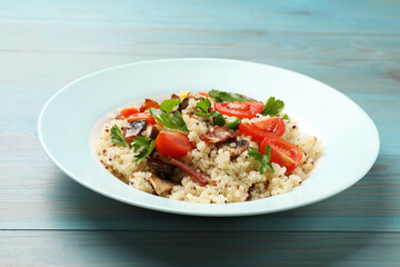 Plate of tasty quinoa porridge with fried bacon, mushrooms and vegetables on light blue wooden table, closeup