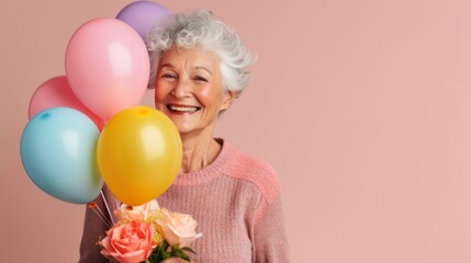 Fototapeta na wymiar Against a clear pink studio backdrop, a senior woman enjoys her birthday with balloons and a special present.