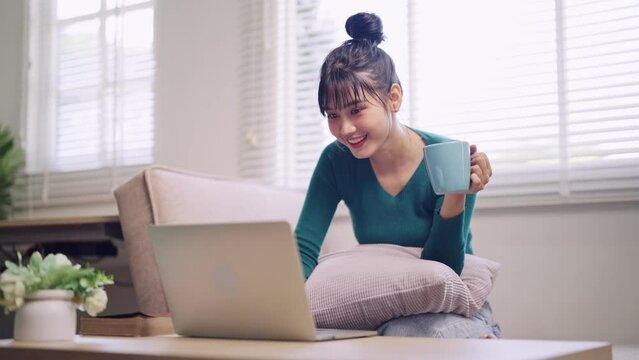 An Asian young woman works comfortably on her laptop, sipping hot coffee while seated on her home sofa during the weekend. Capture the cozy blend of productivity and relaxation.