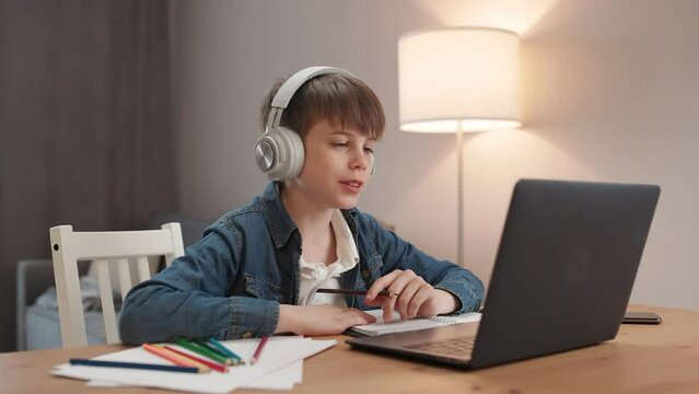 Distance learning by video call, teen boy sitting in a living room and uses a laptop to study in self-isolation at home, the boy in a headphones communicates online.