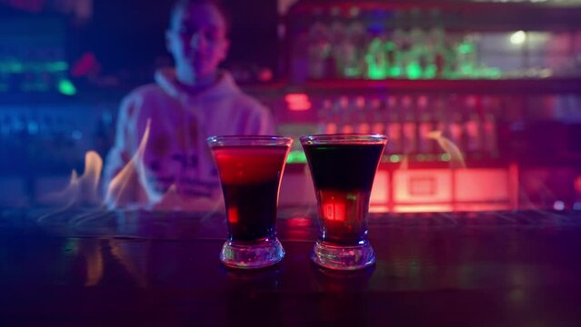 Glass of Alcoholic Drink on a Bar Counter Close-up. Bartender Prepares a Drink by Setting a Glass of Cocktail on Fire in a Nightclub. Bar in Neon Lighting. Slow Motion.
