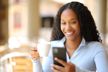 Happy black woman drinking using phone in terrace