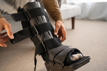 Close-up of Injury man with black splint on leg sitting on the sofa at home and wearing his orthosis. Ankle foot injuries orthosis