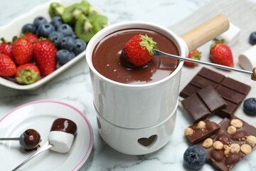 Dipping fresh strawberry in fondue pot with melted chocolate at white marble table