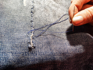 Woman repairing a pair of blue jeans. Fingers with a needle and torn jeans fabric. Sewing up a tear...