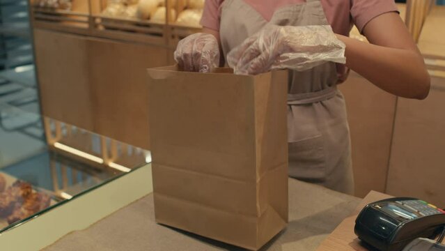 Tilt down shot of female worker in disposable gloves packing fresh bread in paper bag for customer at counter in bakery
