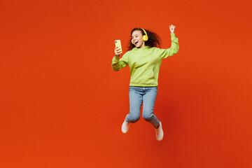 Full body young woman of African American ethnicity wear hoody casual clothes jump high hold use mobile cell phone listen music in headphones do winner gesture isolated on plain red orange background.