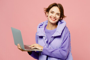 Young smiling happy IT woman she wearing purple vest sweatshirt casual clothes hold use working on laptop pc computer isolated on plain pastel light pink background studio portrait. Lifestyle concept.