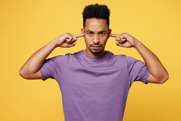 Young man of African American ethnicity he wears purple t-shirt casual clothes cover ears with hands fingers do not want to listen scream isolated on plain yellow background studio. Lifestyle concept.