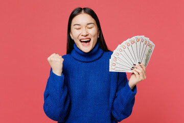 Young woman of Asian ethnicity wear blue sweater casual clothes hold in hand fan of cash money in dollar banknotes do winner gesture isolated on plain pastel light pink background. Lifestyle concept.