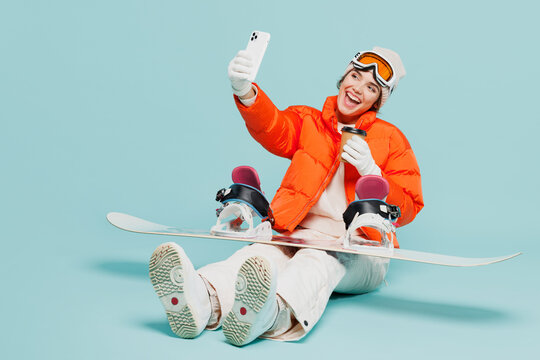 Full body young woman in warm windbreaker jacket ski goggles mask sit with snowboard do selfie mobile cell phone travel rest spend weekend winter season in mountains isolated on plain blue background.