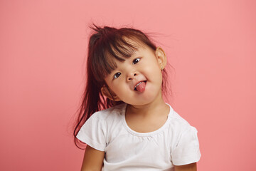 headshot portrait of playful female child tilted her head and shows her tongue standing over a pink...