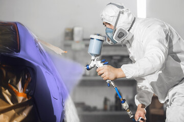 Male worker in protective clothes and mask painting car body to blue violet color using spray paint gun.  - obrazy, fototapety, plakaty