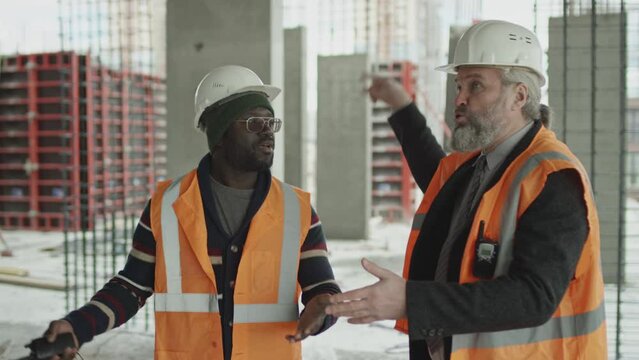 Medium shot of two ethnically diverse emotional engineers in hard hats and safety vests standing at construction site and having heated argument about work process