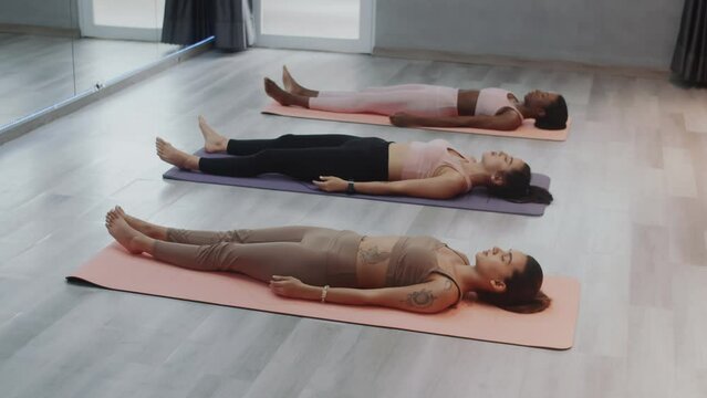 High angle view of group of multi-ethnic female yogis meditating on mats on floor in corps pose while relaxing after stretching