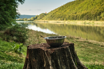 Breakfast by the river. An old stump on the riverbank with a metal bowl with a spoon.