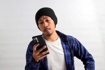 An annoyed young Asian trader is seen looking at his phone with a displeased expression, likely due to losses in the stock market, standing against white background. Finance, and stock market concept