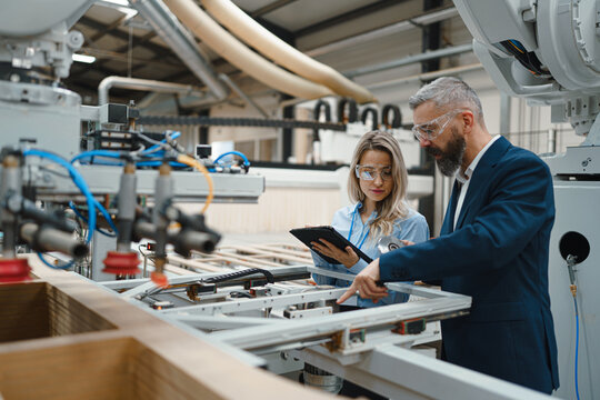 Female engineer and male project manager standing in modern industrial factory, talking about production of wooden furniture. Big furniture manufacturing facility with robotics and automation.