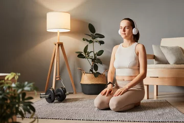 Fotobehang Athletic recovery at home. Sporty floor harmony. Pretty woman wearing beige sportswear relaxing after work out training sitting on floor in living room in headphone listening meditation © sementsova321
