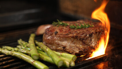 The prefect mouth watering bone-in rib-eye steak cooking on the bbq, barbecue, barbeque or griller with flames and asparagus sides. Rosemary garnish and close-up angle. - obrazy, fototapety, plakaty
