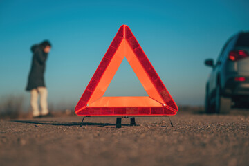 Vehicle breakdown, red warning triangle on the road with a broken down car and woman making a phone call to road service