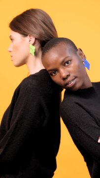 Side view of diverse young female friends leaning on each other looking at camera while wearing trendy colorful earrings against yellow background