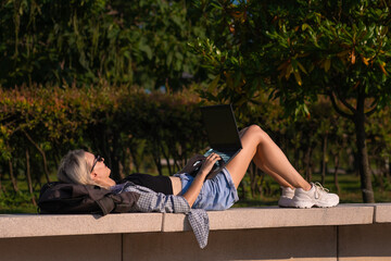 Young slender blonde woman is lying on a park bench with a laptop on a sunny day, side view. Happy girl with a laptop in the park